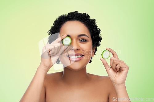 Image of happy african woman making eye mask of cucumbers