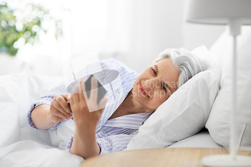 Image of happy senior woman with smartphone in bed at home