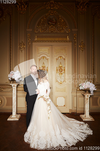Image of Luxury wedding couple in love. Beautiful bride in white dress with brides bouquet and handsome groom in black suit standing in baroque interior and embracing each other