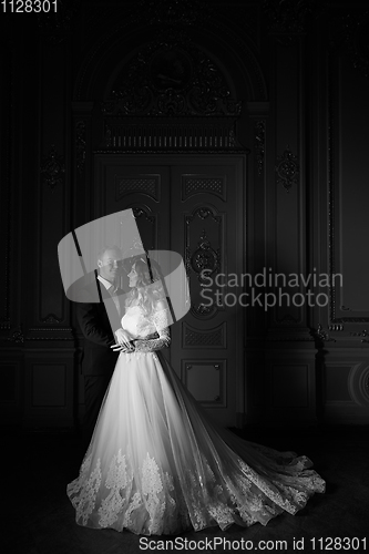 Image of Black and white photography. Luxury wedding couple in love. Beautiful bride in white dress with brides bouquet and handsome groom in black suit standing in baroque interior and embracing each other