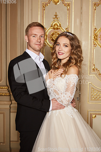 Image of Luxury wedding couple in love. Beautiful bride in white dress with brides bouquet and handsome groom in black suit standing in baroque interior and embracing each other