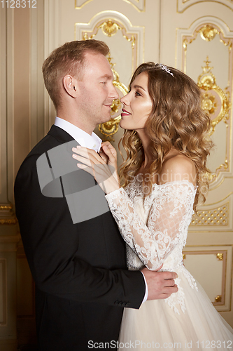 Image of Luxury wedding couple in love. Beautiful bride in white dress with brides bouquet and handsome groom in black suit standing in baroque interior and embracing each other