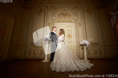 Image of Luxury wedding couple in love. Beautiful bride in white dress with brides bouquet and handsome groom in black suit standing in baroque interior and embracing each other