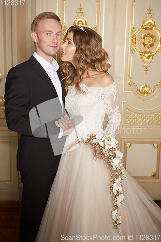 Image of Luxury wedding couple in love. Beautiful bride in white dress with brides bouquet and handsome groom in black suit standing in baroque interior and embracing each other