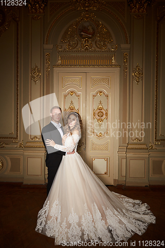 Image of Luxury wedding couple in love. Beautiful bride in white dress with brides bouquet and handsome groom in black suit standing in baroque interior and embracing each other