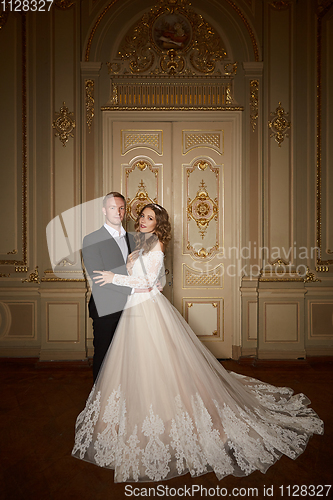 Image of Luxury wedding couple in love. Beautiful bride in white dress with brides bouquet and handsome groom in black suit standing in baroque interior and embracing each other