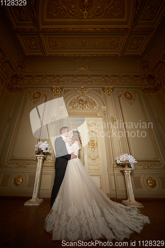 Image of Luxury wedding couple in love. Beautiful bride in white dress with brides bouquet and handsome groom in black suit standing in baroque interior and embracing each other