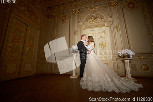 Image of Luxury wedding couple in love. Beautiful bride in white dress with brides bouquet and handsome groom in black suit standing in baroque interior and embracing each other