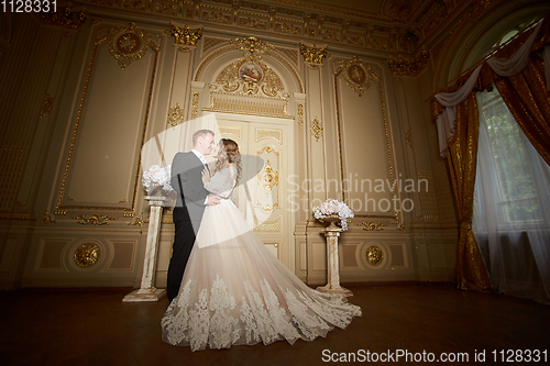 Image of Luxury wedding couple in love. Beautiful bride in white dress with brides bouquet and handsome groom in black suit standing in baroque interior and embracing each other