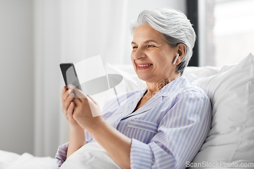 Image of senior woman with smartphone and earphones in bed