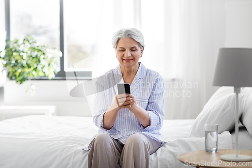 Image of happy senior woman with smartphone on bed at home