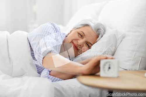 Image of happy senior woman with alarm clock in bed at home