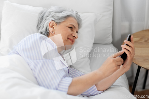 Image of happy senior woman with smartphone in bed at home