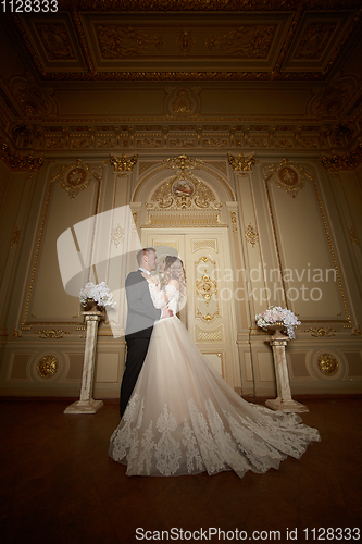 Image of Luxury wedding couple in love. Beautiful bride in white dress with brides bouquet and handsome groom in black suit standing in baroque interior and embracing each other