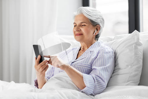 Image of senior woman with smartphone and earphones in bed
