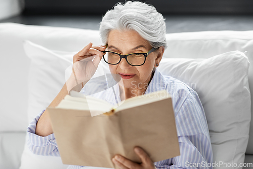 Image of old woman in glasses reading book in bed at home