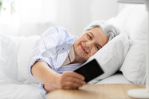 Image of happy senior woman with smartphone in bed at home