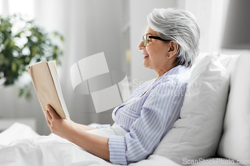 Image of old woman in glasses reading book in bed at home