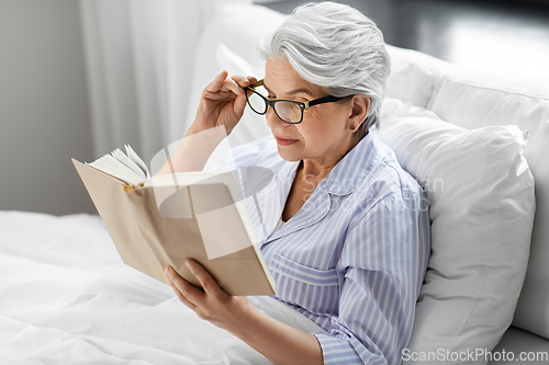 Image of old woman in glasses reading book in bed at home