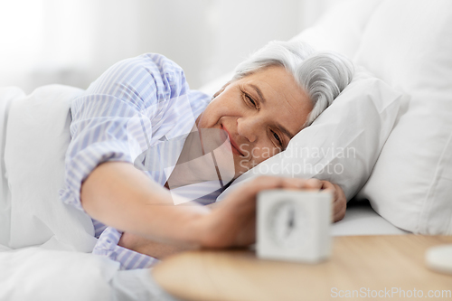 Image of happy senior woman with alarm clock in bed at home