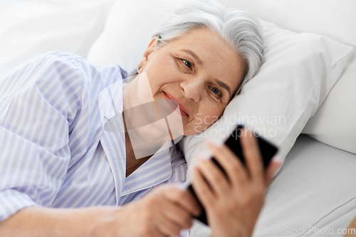 Image of happy senior woman with smartphone in bed at home