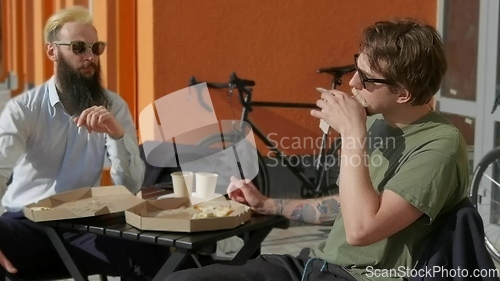 Image of Friendly talk. Two happy young men eating pizza and talking to each other while sitting outdoors