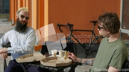 Image of Friendly talk. Two happy young men eating pizza and talking to each other while sitting outdoors.