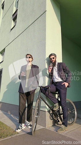 Image of Two young men friends dressed casually spending time together at the city, drinking takeaway coffee next bike.