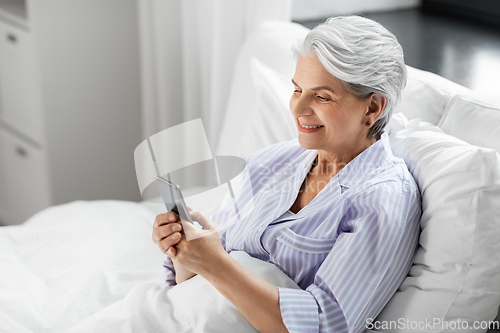 Image of happy senior woman using smartphone in bed at home