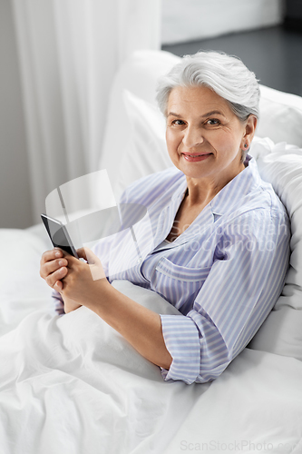 Image of happy senior woman using smartphone in bed at home