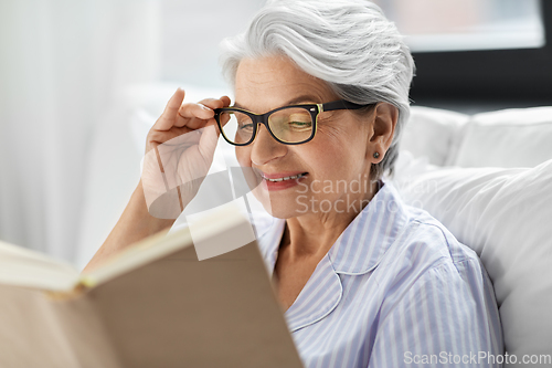 Image of old woman in glasses reading book in bed at home