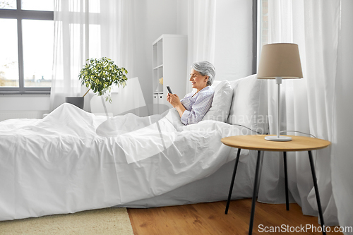 Image of happy senior woman using smartphone in bed at home