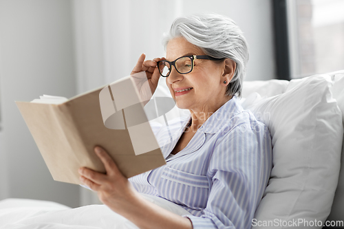 Image of old woman in glasses reading book in bed at home