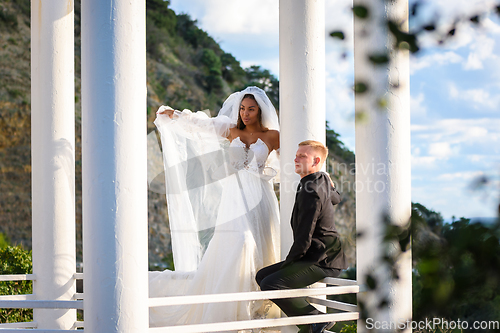 Image of The newlyweds are walking in the gazebo, the girl is standing with her veil raised, the guy is sitting on the railing