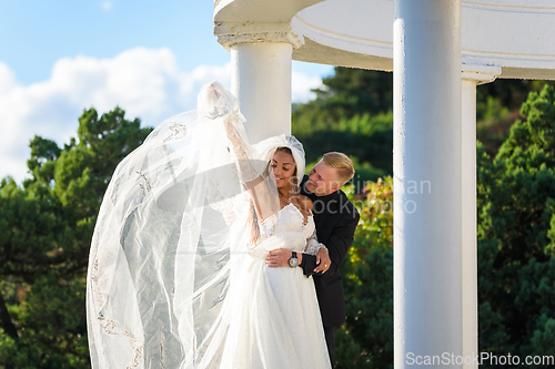 Image of The newlyweds happily embrace in a beautiful gazebo against the backdrop of foliage and sky