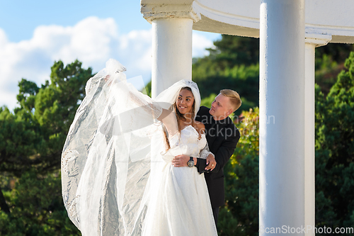 Image of Happy newlyweds hugging on a walk, the girl raised her veil with her hand and smiles