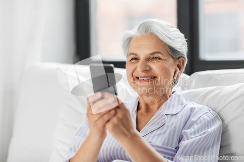 Image of senior woman with smartphone and earphones in bed