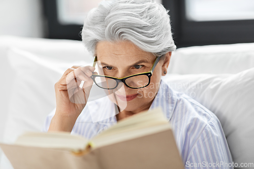 Image of old woman in glasses reading book in bed at home