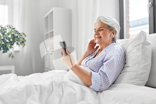 Image of senior woman with smartphone and earphones in bed