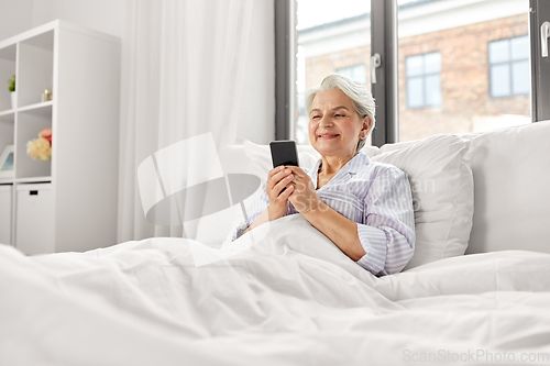 Image of happy senior woman using smartphone in bed at home