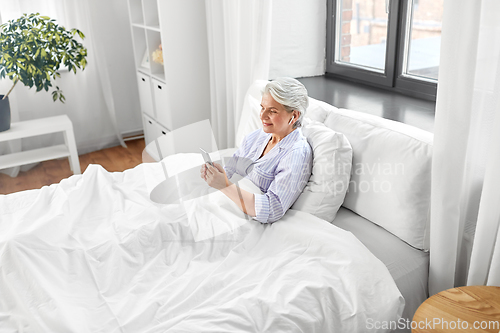 Image of senior woman with smartphone and earphones in bed