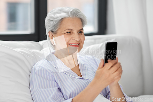 Image of senior woman with smartphone and earphones in bed