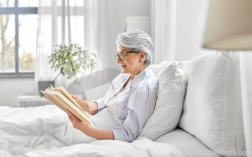 Image of old woman in glasses reading book in bed at home