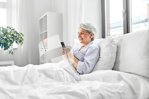 Image of happy senior woman using smartphone in bed at home