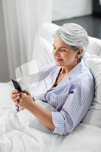 Image of happy senior woman using smartphone in bed at home