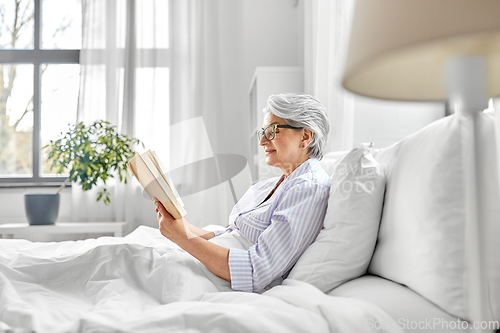 Image of old woman in glasses reading book in bed at home