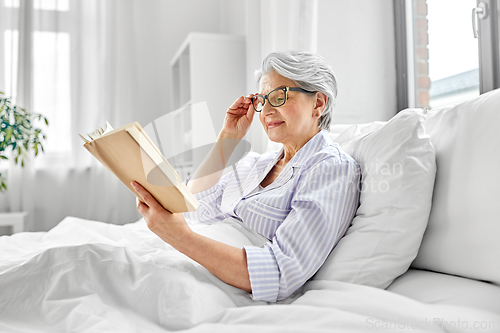 Image of old woman in glasses reading book in bed at home