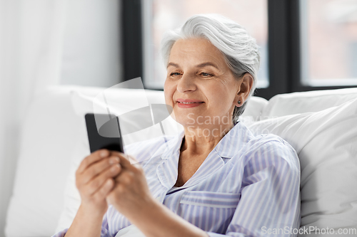 Image of happy senior woman using smartphone in bed at home