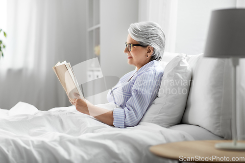 Image of old woman in glasses reading book in bed at home
