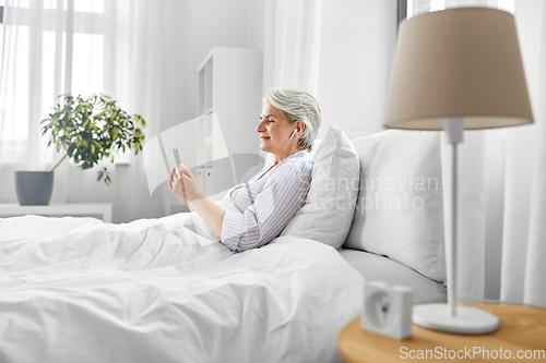 Image of senior woman with smartphone and earphones in bed